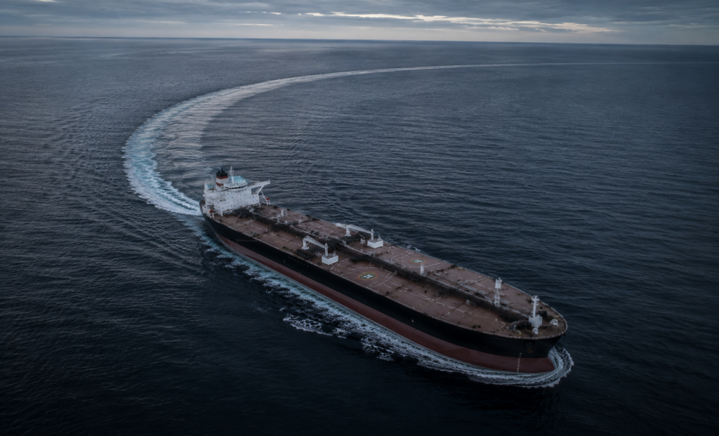 Aerial view of a large commercial tanker making a gradual turn through dark open water, leaving a broad curved wake behind it